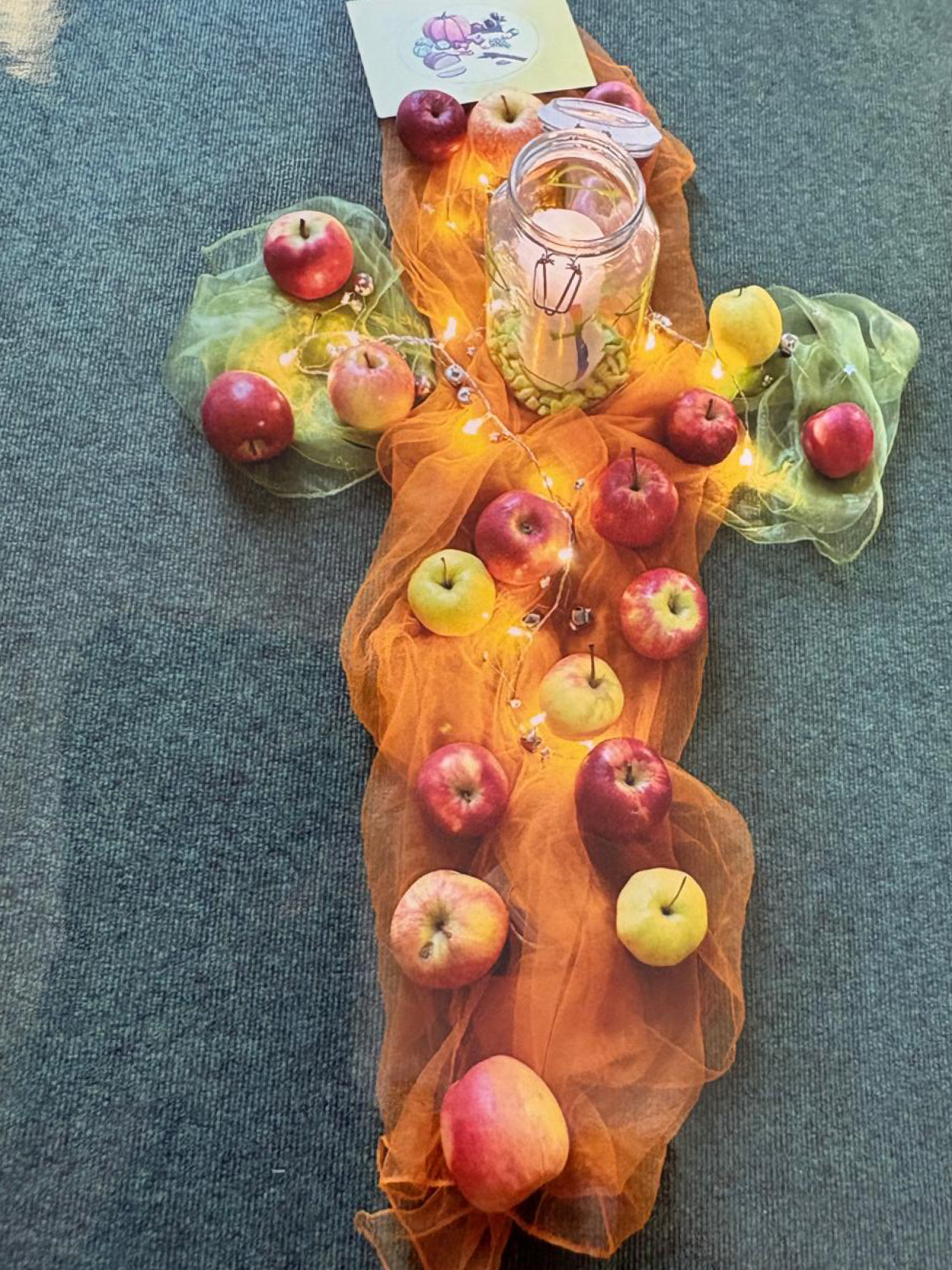 Ein auf dem Boden gestaltetes Kreuz aus orangefarbenen und grünen Tüchern, dekoriert mit vielen Äpfeln, einer Lichterkette und einem Windlichtglas.
