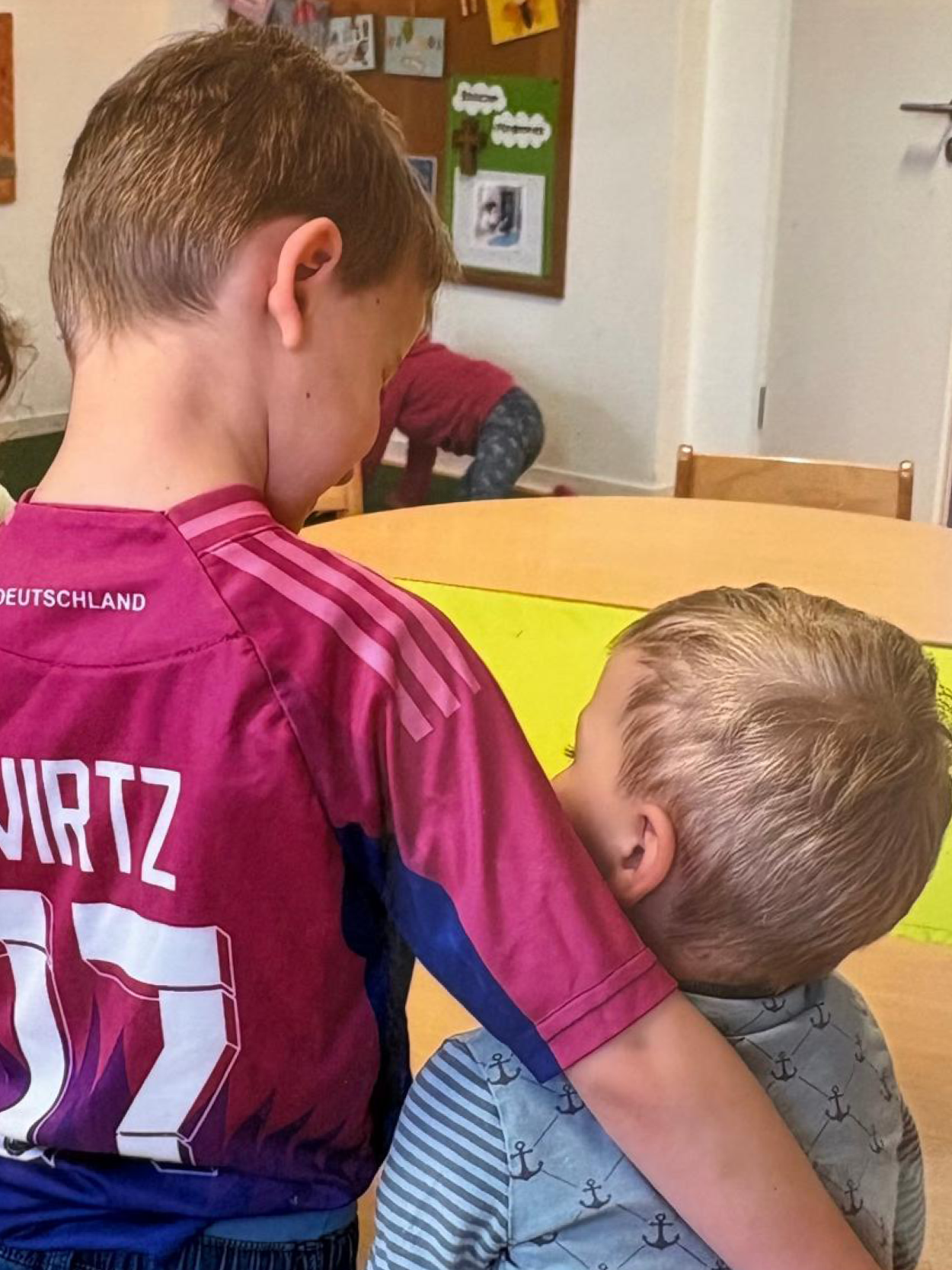 Ein Junge im Trikot nimmt ein anderes Kind im Gruppenraum freundschaftlich in den Arm.
